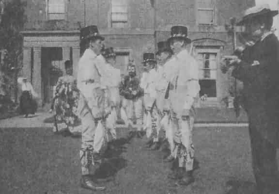 THE BIDFORD MORRIS SIDE.<br /> (FIDDLER in foreground, to the right; HOBBY-HORSE—left, and FOOL—centre, beyond Dancers.)