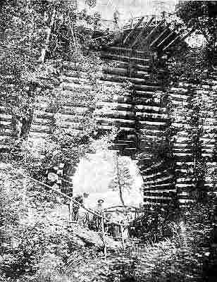 Tall horizontal log bridge; wooden stair rail leading to men working in gulch below.